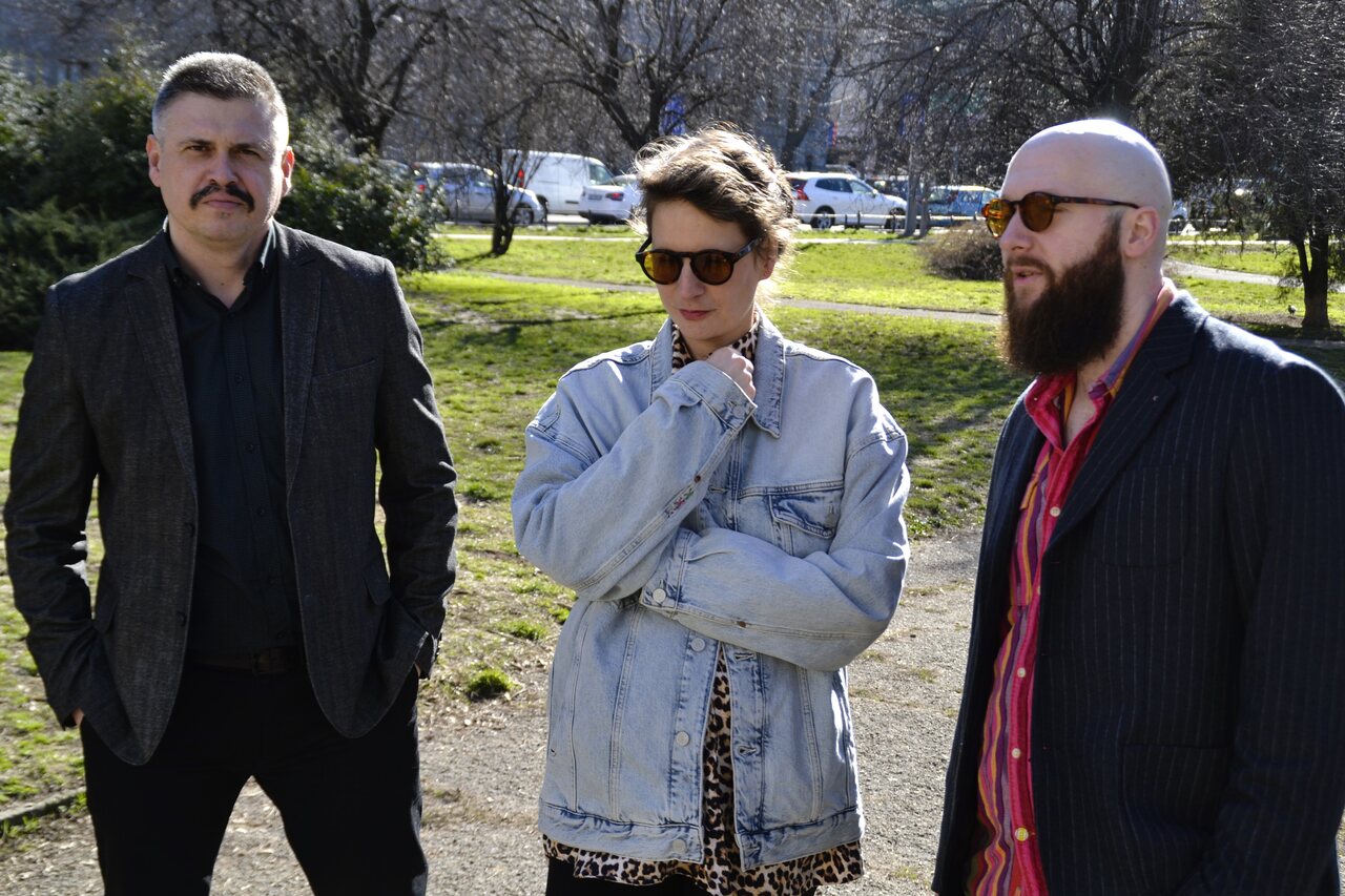 The three founders standing in a park — two in dark jackets and one in a denim jacket